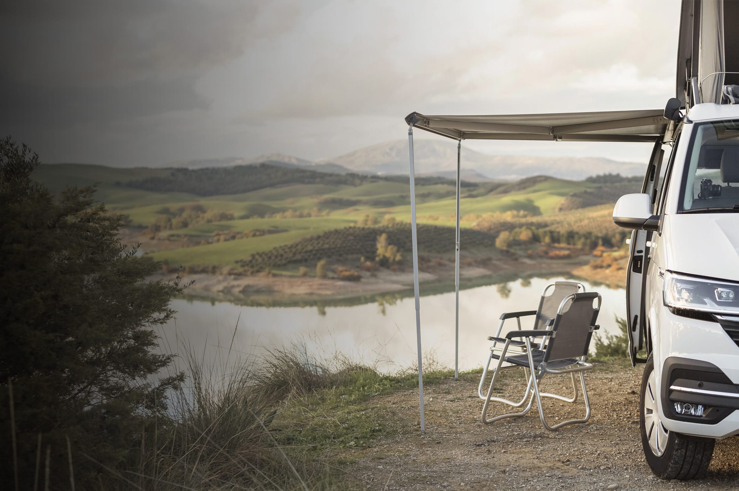Ein VW California steht mir ausgefahrener Markise und zwei Campingstühlen zum Mieten vor einer traumhaften Landschaft.