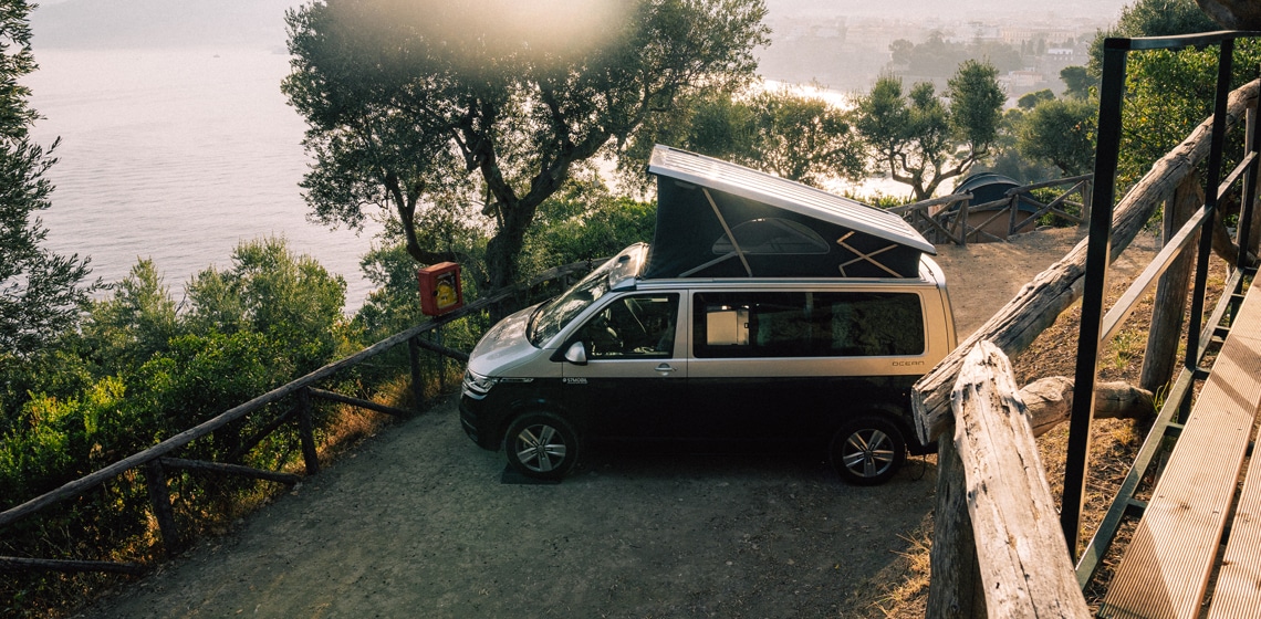 Ein VW California mit Aufstelldach auf einem Campingplatz am Wasser geparkt, mit einem Sonnenuntergang im Hintergrund - perfekt zum Mieten geeignet.