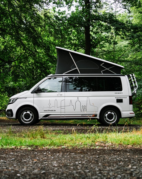 Seitenansicht eines weißen VW Californias mit praktischem Aufstelldach, Gepäckträger und Aufdruck der Siegener Skyline - zum Mieten erhältlich!